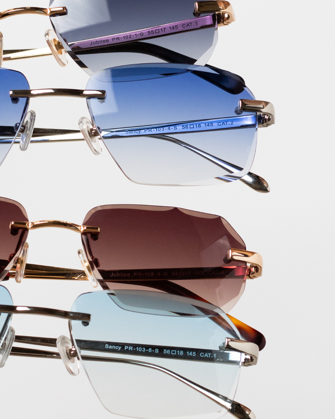 An artistic stack of four luxury sunglasses from different collections, each lens displaying a unique gradient. From top to bottom: 'Jubilee' with gradient grey lenses in 18k gold frames, 'Sancy' in onyx blue with 18k gold, another 'Jubilee' pair in modern brown, and 'Sancy' with sky blue lenses in sterling silver frames, all with a signature diamond cut. The reflective quality of the lenses shines against the white backdrop, showcasing the brand's diverse yet consistently elegant style.