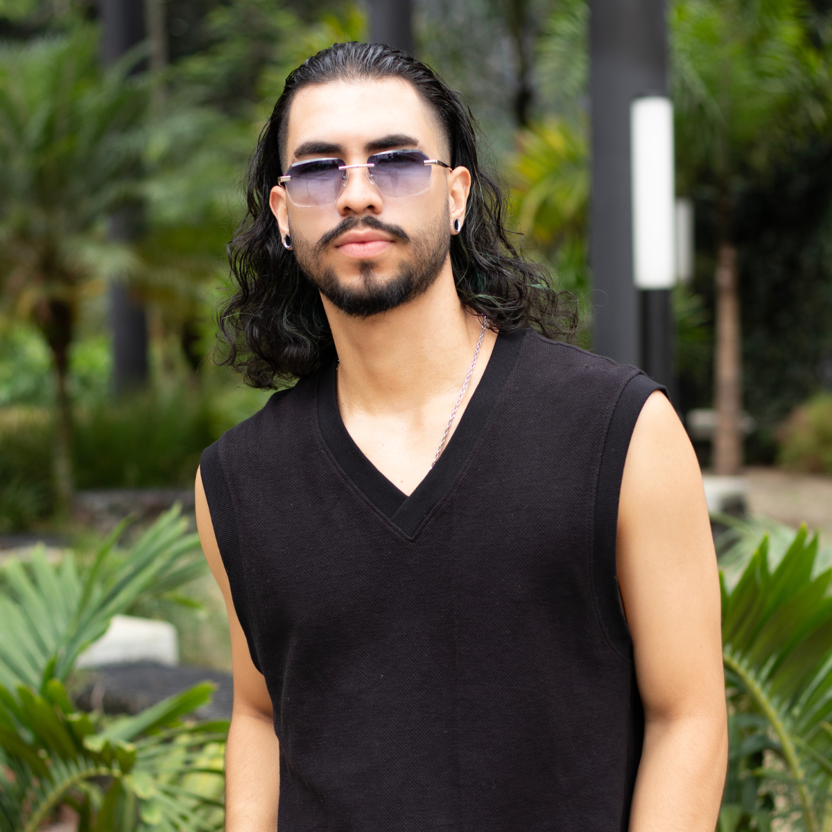 A model confidently wears 'Sancy' rimless sunglasses in gradient grey. The photo captures him in a casual black V-neck, with the rimless glasses complementing his relaxed yet stylish look. The lush greenery in the background provides a natural contrast that underscores the sunglasses' chic and versatile design.