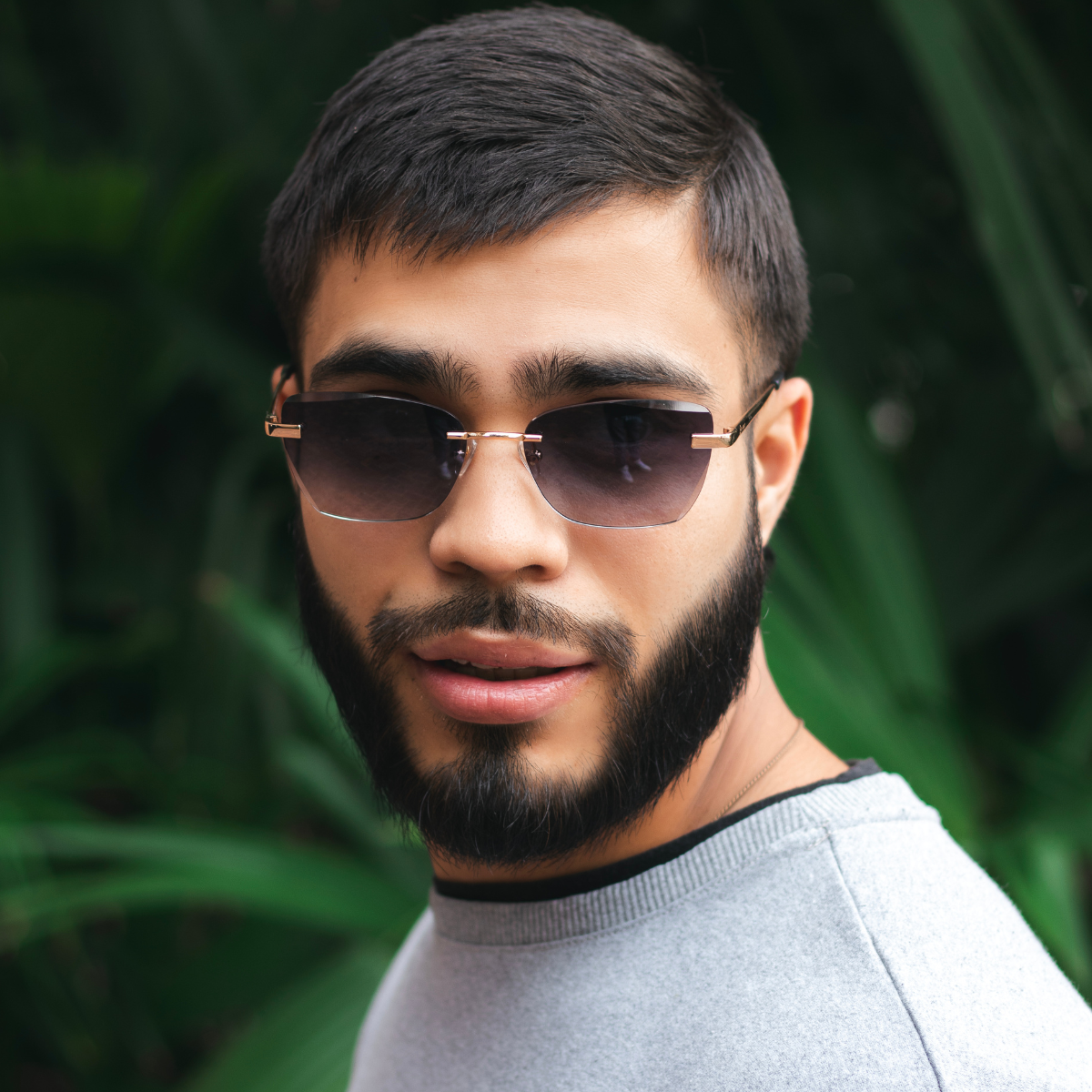 rey. The 18k gold-plated frames with diamond cut detailing catch the light, complementing his stylish beard and contemporary attire. Lush foliage softly blurs in the background, emphasizing the luxurious and fashion-forward design of the rimless eyewear.