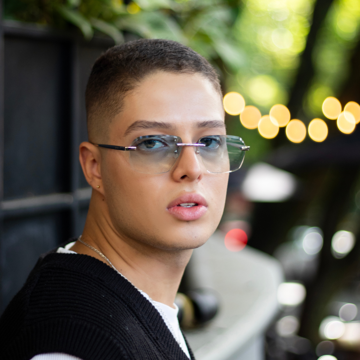 A close-up of a model wearing 'Sancy' rimless sunglasses with sky blue lenses. The rimless design complements the model's modern style against a blurred natural background, accentuated by warm bokeh lights that highlight the eyewear's elegant and contemporary aesthetic