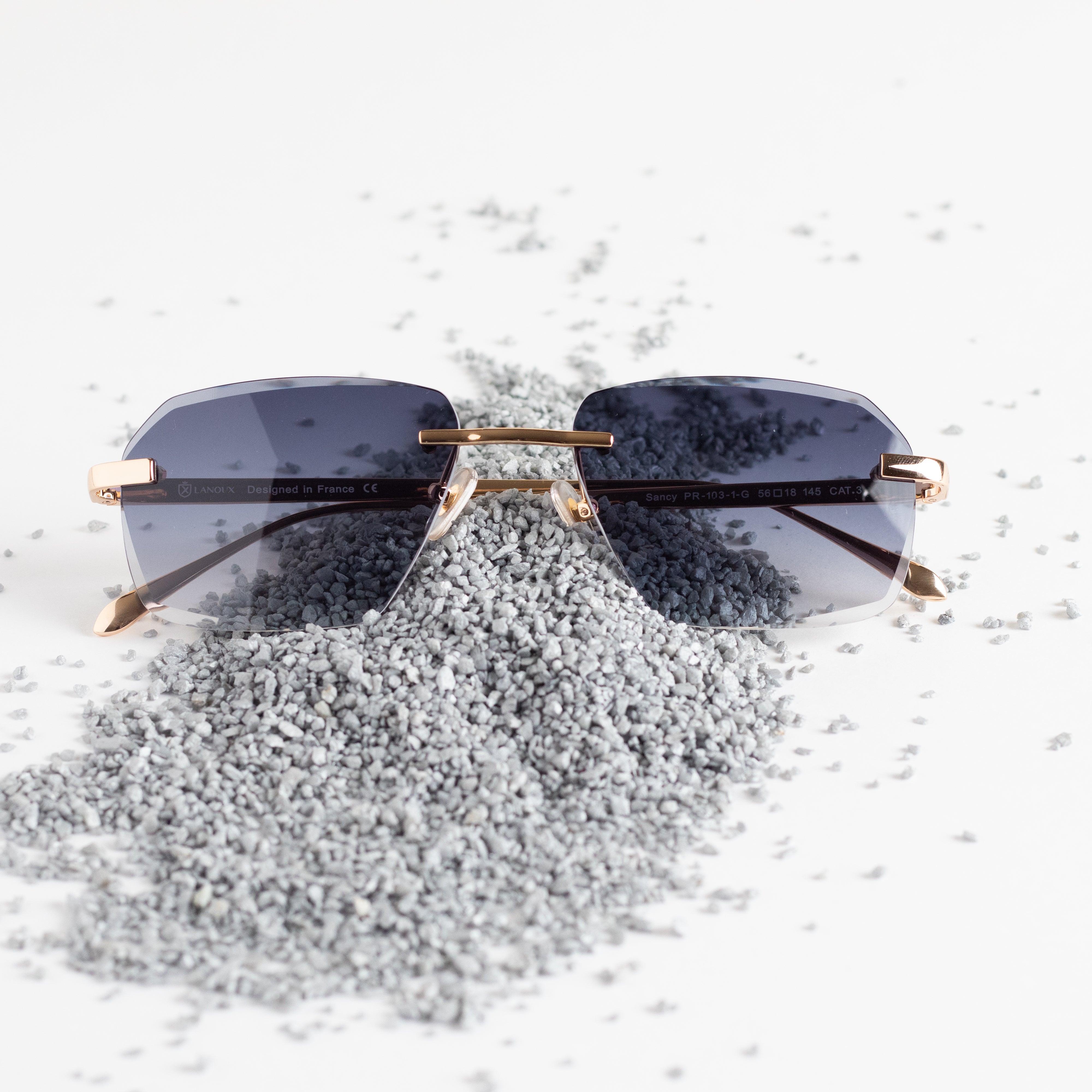 he 'Sancy' sunglasses, poised on a speckled grey surface, feature gradient grey lenses and illustrious 18k gold frames with a diamond cut finish. The refined aesthetic of the glasses is accentuated by the contrasting granular texture beneath them, illustrating the fusion of craftsmanship and sophisticated style.
