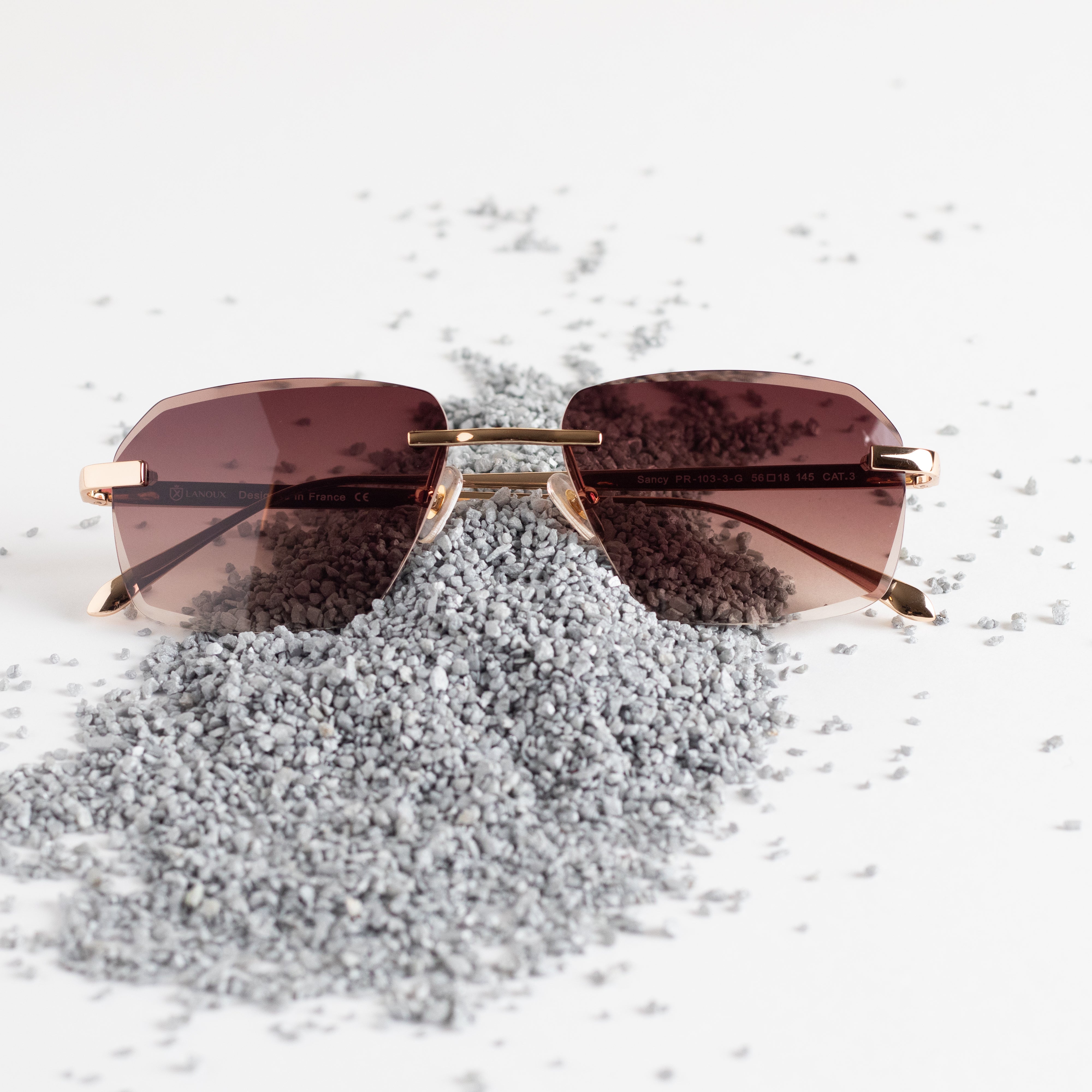 The 'Sancy' rimless sunglasses are displayed on a textured surface, their modern brown lenses complementing the 18k gold frames with a diamond cut. The elegant design and rich color palette are highlighted against a backdrop of scattered grey granules, symbolizing the brand's attention to detail and luxury.